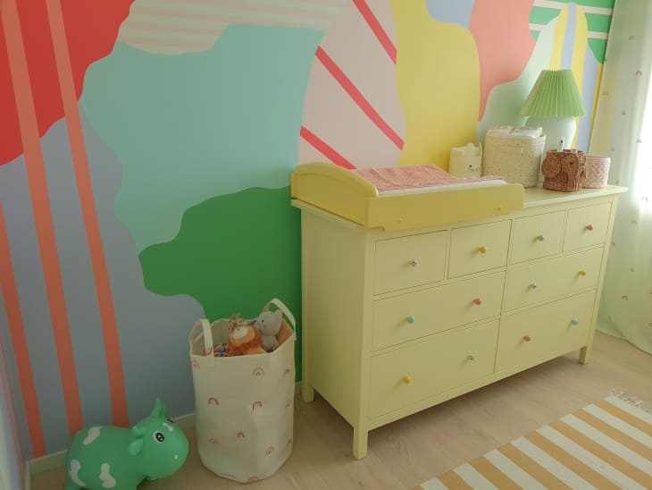 multi colored splatter wall mural with stripes throughout, yellow dresser, yellow changing table on top, toys, basket of toys, light airy white curtains, green pleated lamp shade, wood floors, yellow and white striped rug