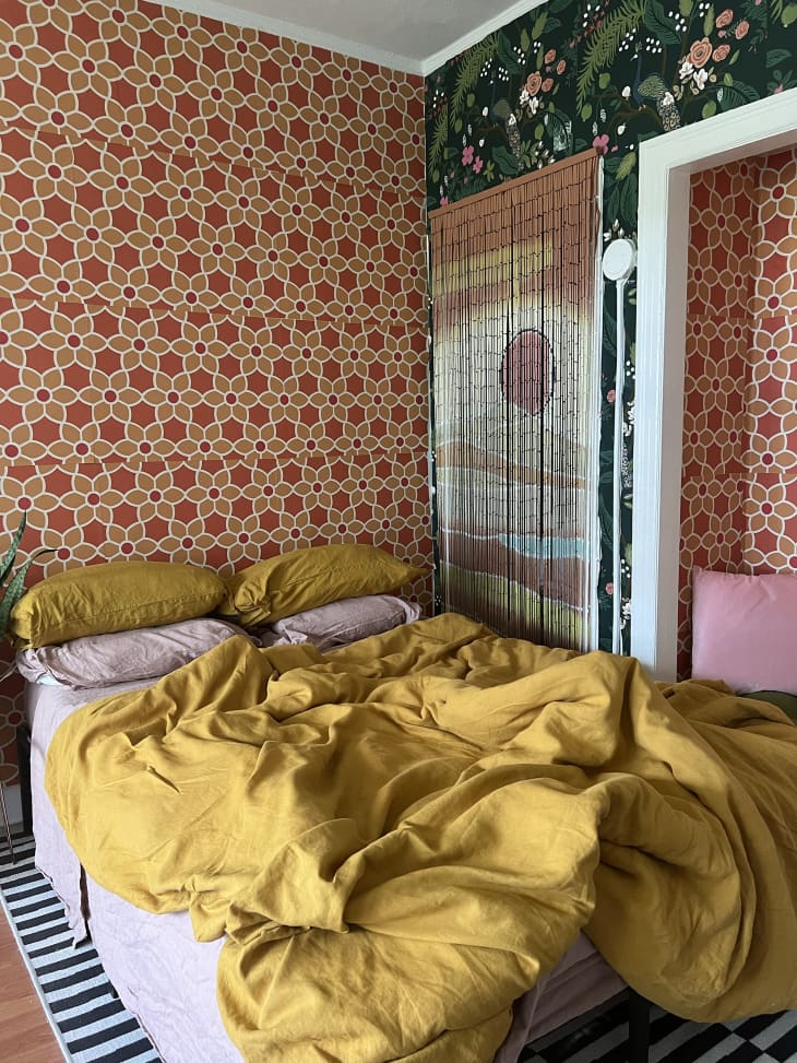 red and white geometric pattern wallpaper, dark floral wallpaper, white door frame, yellow comforter, white sheets, yellow pillow cases, bead curtain, black and white striped rug, wall cut out, pink lamp shade, wood floors