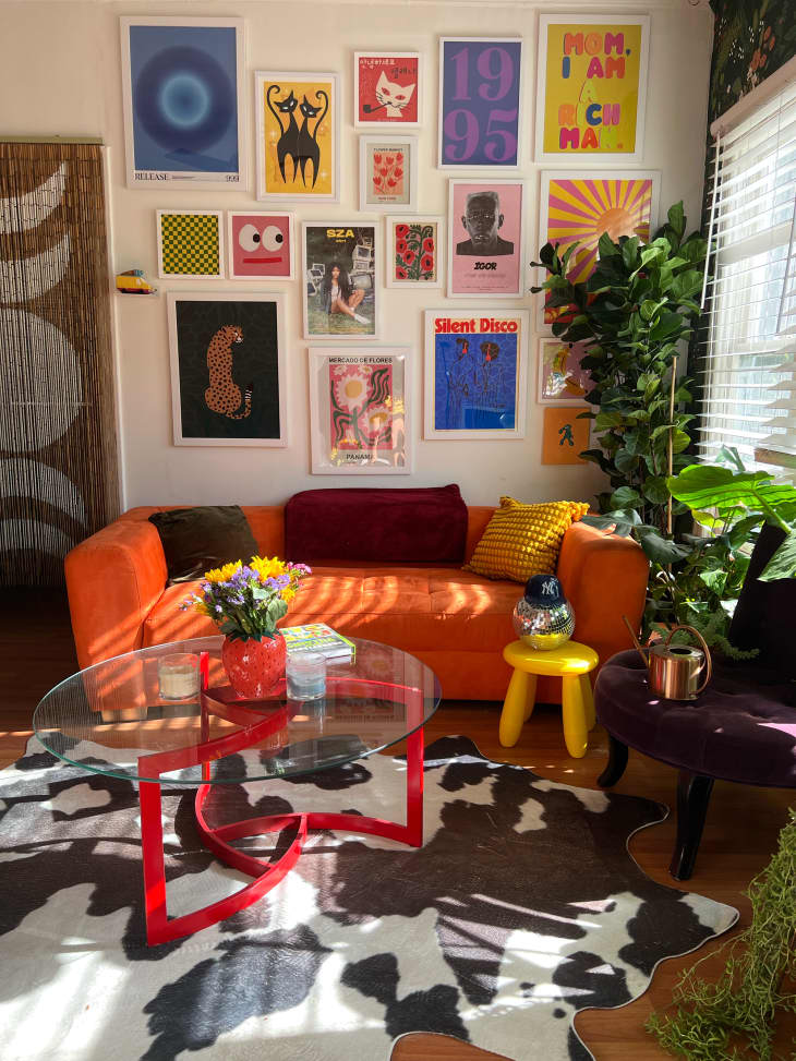 gallery wall, graphic art, orange fabric couch, curved couch, cow print rug, beads in door frame, plants, yellow tri leg bubble stool, flowers, deep purple fabric bench, red leg coffee table with glass top, disco ball, wood floors