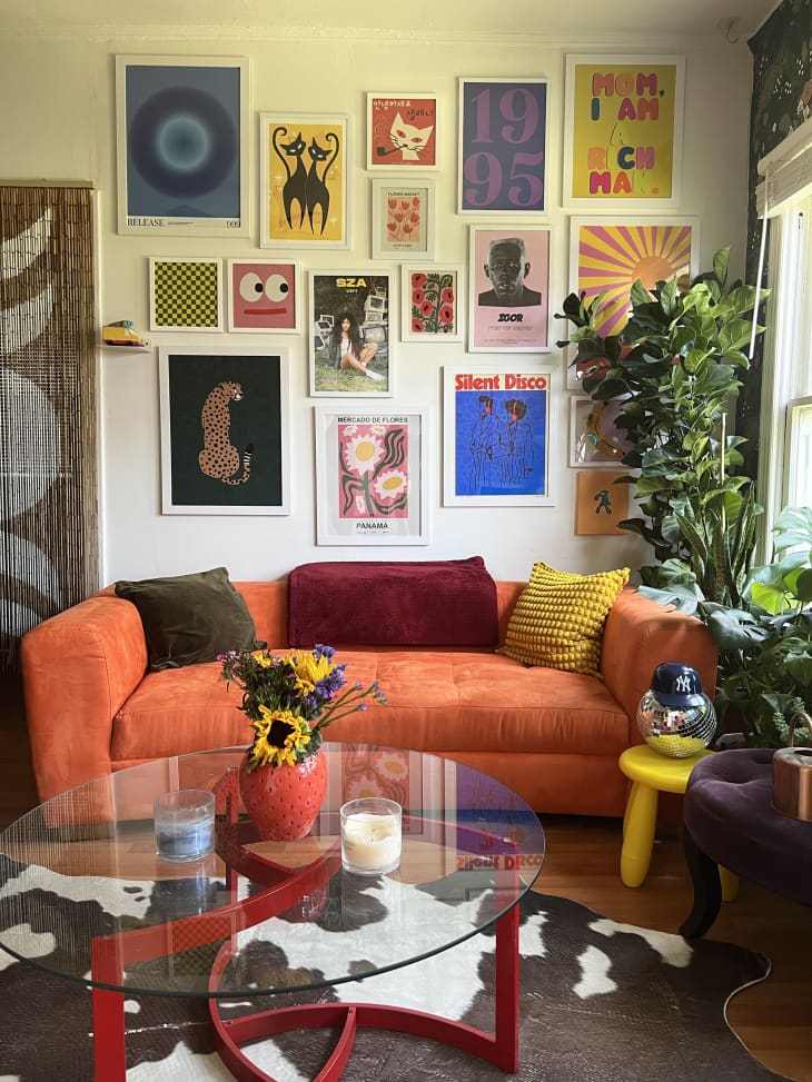 gallery wall, graphic art, orange fabric couch, curved couch, cow print rug, beads in door frame, plants, yellow tri leg bubble stool, flowers, deep purple fabric bench, red leg coffee table with glass top, disco ball, wood floors