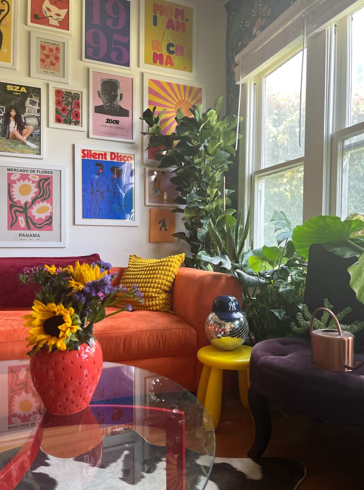 gallery wall, graphic art, orange fabric couch, curved couch, cow print rug,  plants, yellow tri leg bubble stool, flowers, deep purple fabric bench, red leg coffee table with glass top, strawberry vase, disco ball, wood floors