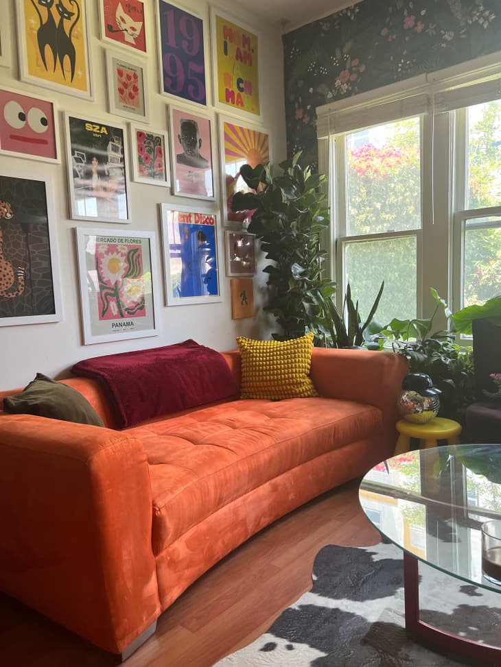 gallery wall, graphic art, orange fabric couch, curved couch, cow print rug, plants, yellow tri leg bubble stool, flowers, deep, red leg coffee table with glass top, disco ball, dark patterned wallpaper accent wall, wood floors