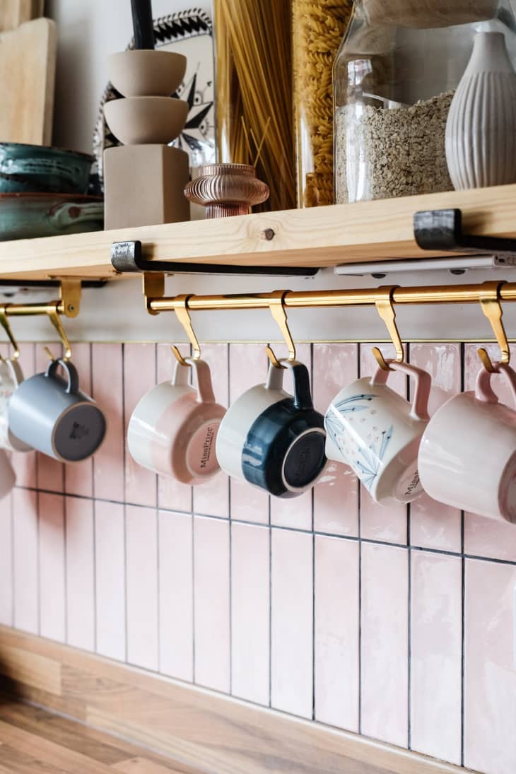 hanging mugs, glossy pink subway tile, floating shelves, dried pasta in glass containers, oats in glass jar, butcher block countertops, small bowls