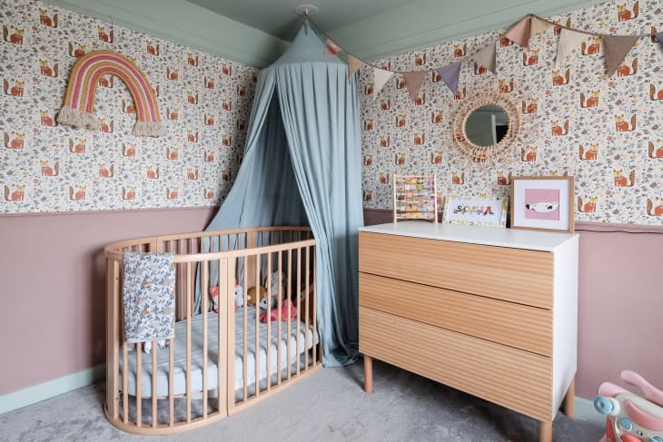 illustrated fox pattern wallpaper on half wall, dusty pink paint on bottom half of wall, blue ceiling, natural wood dresser with white top and side, triangle flag streamer, beige carpet, baby blue curtains above crib, circle mirror, rainbow fibers art, oval crib with wood slates