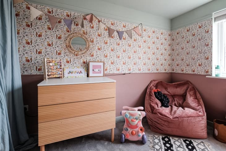 illustrated fox pattern wallpaper on half wall, dusty pink paint on bottom half of wall, dusty pink bean bag chair, blue ceiling, natural wood dresser with white top, triangle flag streamer, beige carpet, baby blue curtains, kids scooter toy, circle mirror