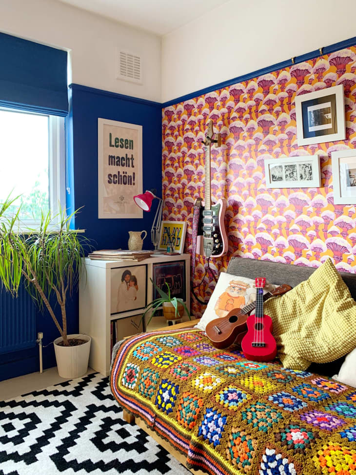 colorful vegetable pattern wallpaper, day bed, granny square blanket, guitars, throw pillows, records, black and white printed rug, plants, black and white art, cardboard box, blue color blocked wall, plants