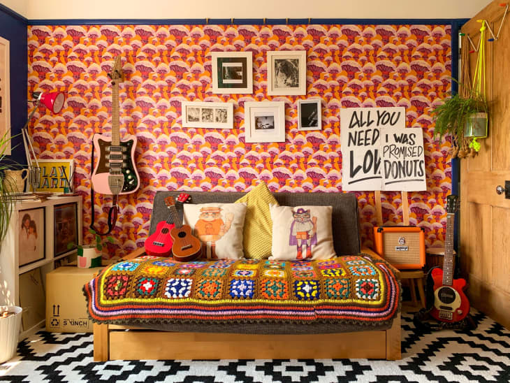 colorful vegetable pattern wallpaper, picket signs, day bed, granny square blanket, guitars, throw pillows, records, black and white printed rug, natural wood door, plants, black and white art, orange amp, cardboard box