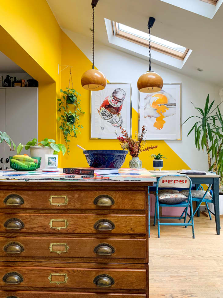 kitchen, plants, colorful patterned counter top, table, pepsi chairs, ketchup art, egg art, round tan lighting fixtures, natural wood, wood floors, skylight, angled ceiling, yellow and white color blocked wall, skinny drawers