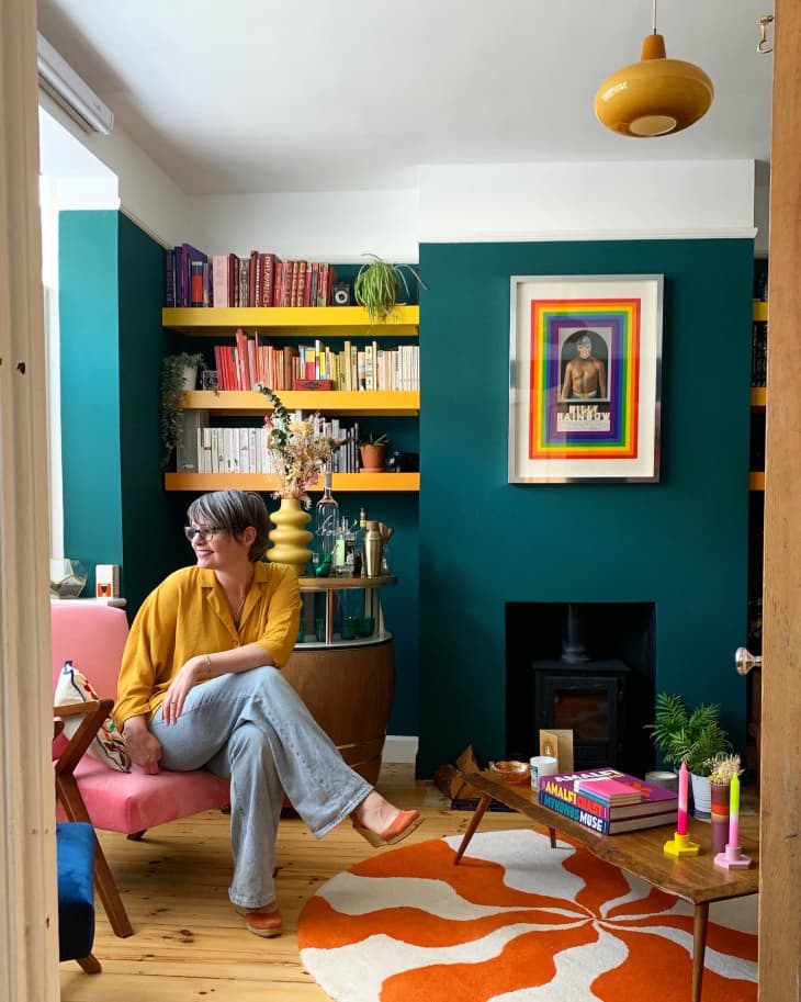 teal wall, open book shelf, yellow book shelf, orange and white wavey circle rug, yellow ronded light fixture, pink arm chair. fireplace, wood floors