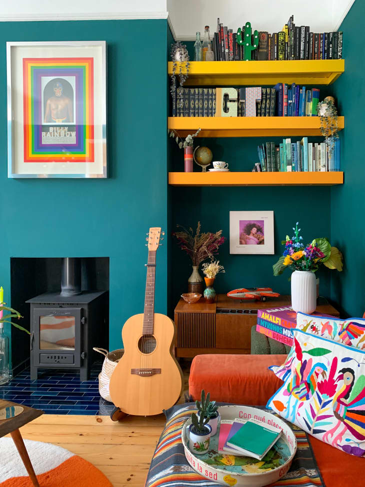 teal wall, open book shelf, yellow book shelf, orange and white wavey circle rug, fireplace, wood floors, plants, guitar, orange couch, colorful throw pillow, colorful throw