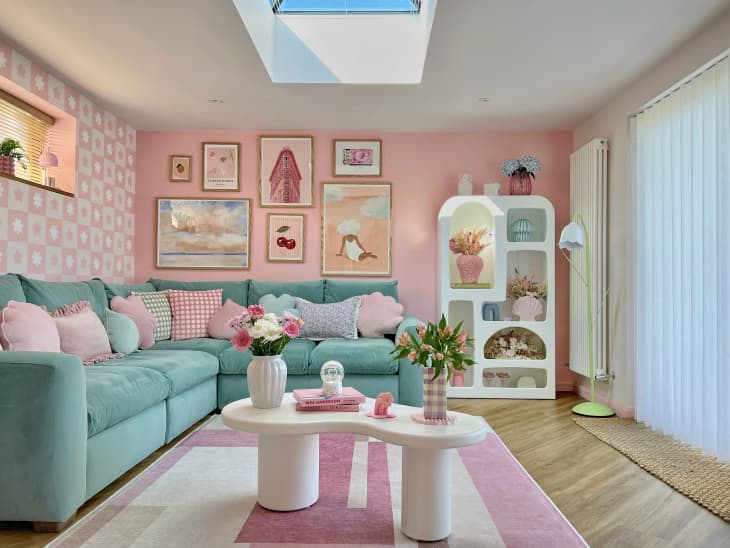 pink walls, curved cut out white book shelf, gallery wall, graphic art, teal soft couch, wood floors, white shag rug, curved white coffee table, pink shag throw, pastel throw pillows, flowers, checkered vase, books, plants, skylight, pink and white geometric rug, pink checkered wall