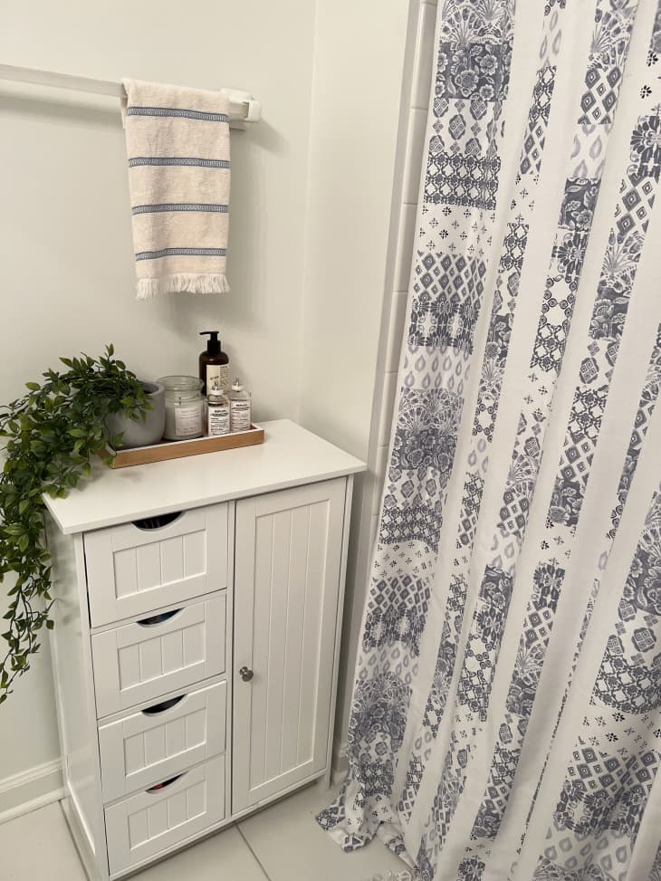 Detail of bathroom with white and blue shower curtain, white cabinet/drawers with tray of products, plant. Blue and white striped towel on bar above