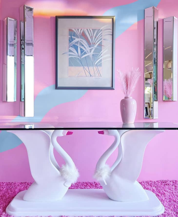 pink wall with blue squiggly line mural, white double swan table base, glass top, skinny mirrors creating design on wall, pink carpet, vintage 80s leaf painting, pink vase