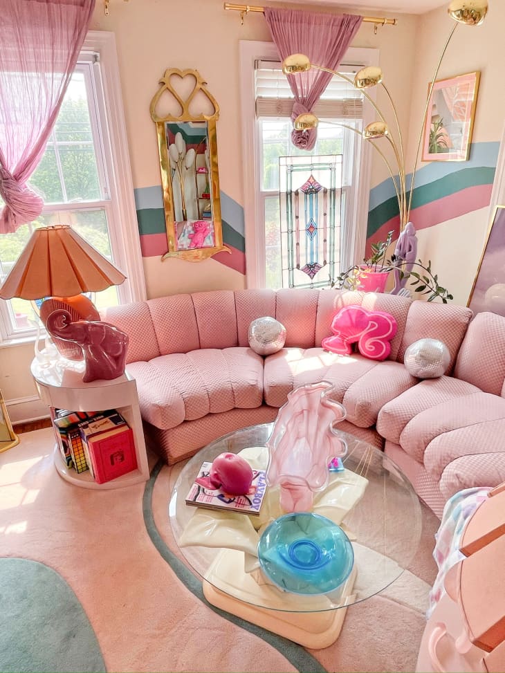 soft pink, striped mural, vintage floor lamp, stained glass window, curved sectional, barbie pillow, blue and pink rug, glass coffee table, light curtains