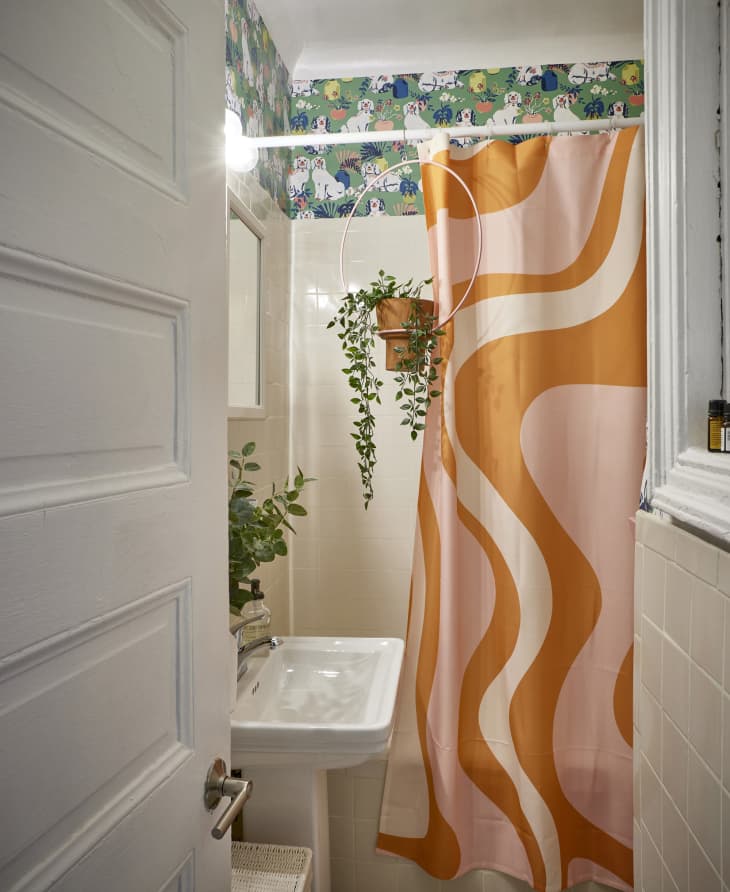 Wavy orange and pink shower curtain in colorful bathroom with green floral wallpaper and a hanging plant in the shower.