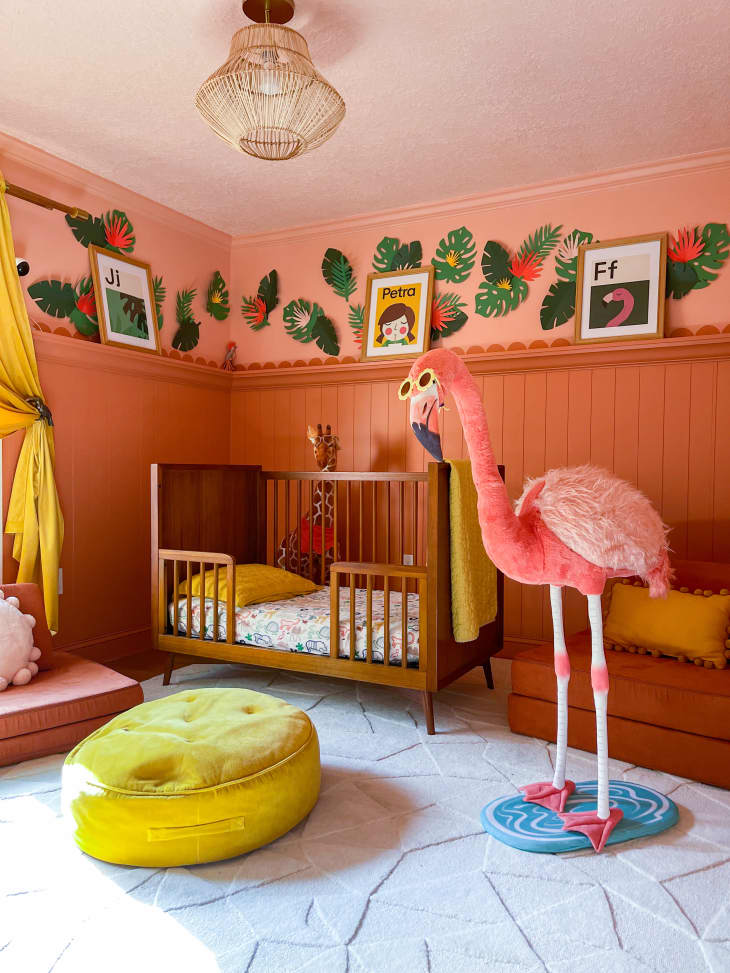 large human life size flamingo statue wearing sunglasses, large round yellow velvet floor pillow, beige patterned rug, 3/4 wall paneling, wall trim, pink and dusty rose walls, dusty rose velvet arm chair, yellow throw pillow, mid century inspired wood crib, small child's bed, picture shelf, leafy mural on wall, yellow curtains, sunlight, rope design chandelier