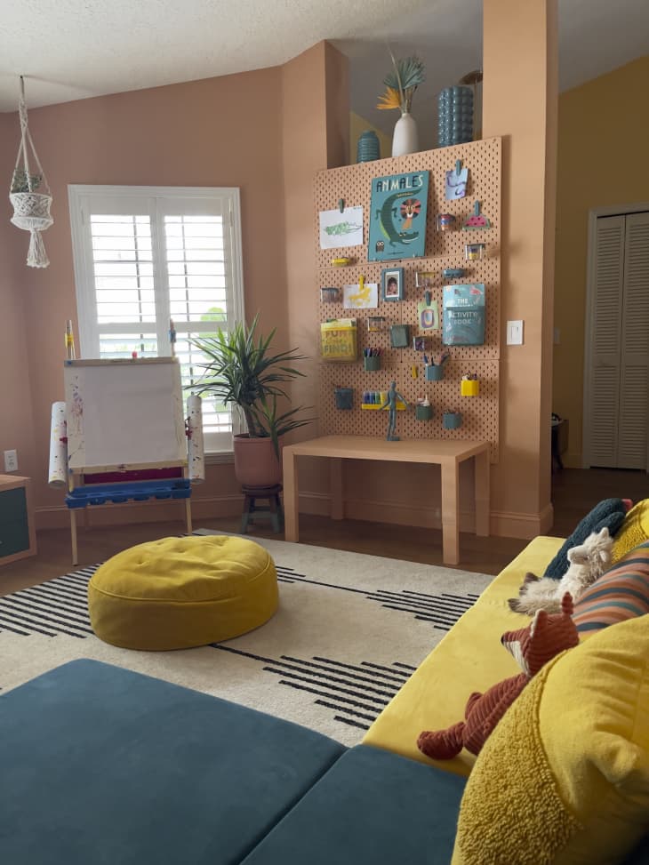 peg board wall, peach, peach table, hanging crafts and kid's art, plant, wood floors, pink plant pot, kid's easel, large yellow floor pillow, yellow and teal foldable couch pillows, cream and black striped geometric rug, hanging plant, wall cut out, angled ceiling