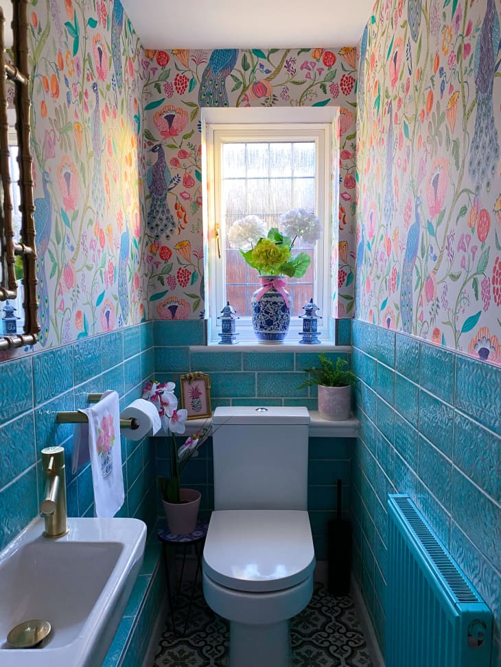 bathroom, teal tile, narrow, colorful peacock patterned wallpaper, plant, half wall wallpaper, white toilet, gold sink fixtures