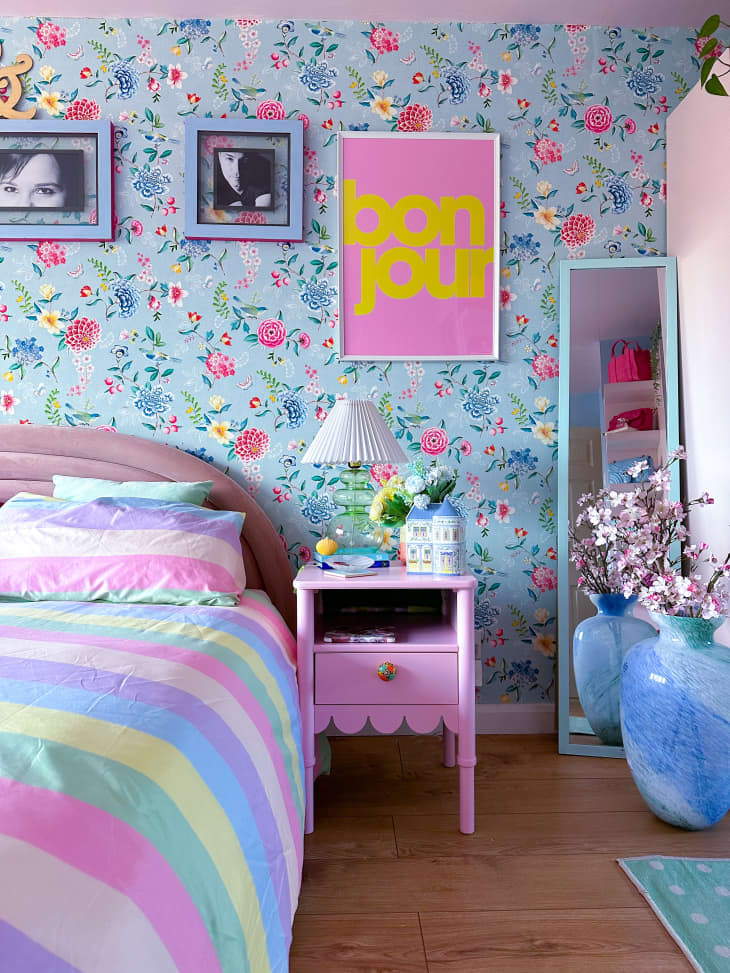 bedroom, blue and pink floral wallpaper, pink ceiling, striped comforter, floor mirror, green rug, hard wood floors, accent pillow, colorful art, plants, pink plush headboard, pink scalloped end table