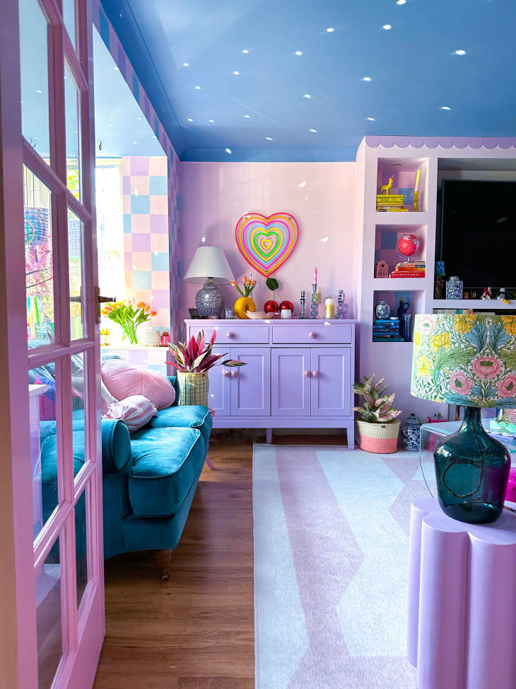 pink door frame, wavy pink stripe walls, purple cabinet, heart art, sunlight, teal velvet loveseat, pink and white rug, hard wood floor, knickknacks, blue ceiling, flower cut out end table, teal lamp, colorful floral patterned lamp shade, tv built in to wall
