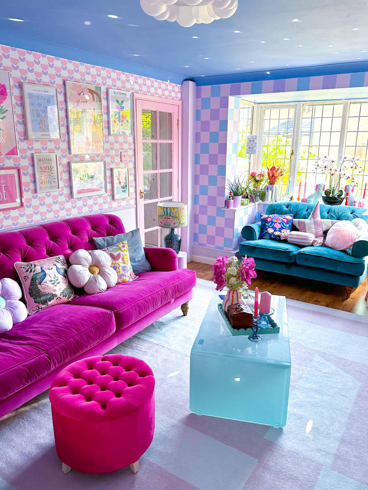 living room, pink velvet couch, teal velvet loveseat, pink velvet ottoman, pink flower wallpaper, pink purple and blue checkered wall, window, blue ceiling, pink and white patterned rug, acrylic waterfall coffee table, accent pillows, gallery wall, pink door frame