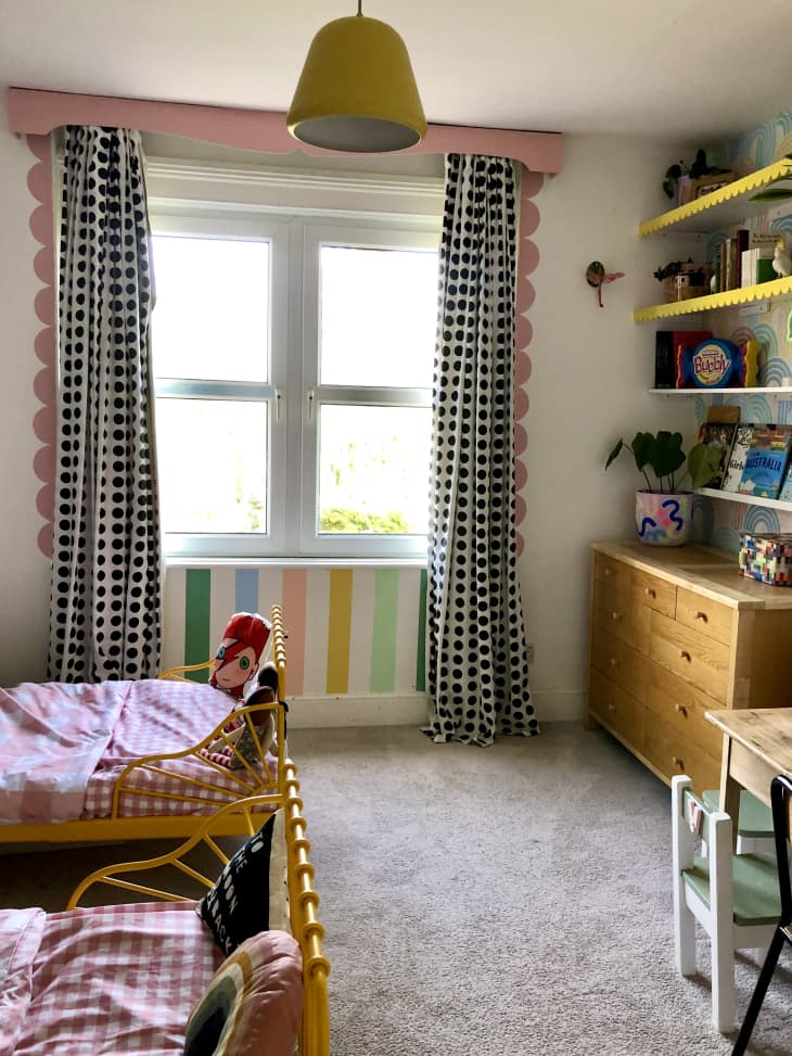 beige carpet, yellow metal bed frames with footers, black and white checkered curtains, pink and white checkered sheets, rainbow stripes below window, pink scallop framed window, pink wooden curtain valance, yellow scalloped trim on floating book shelves, wooden dresser, small white child's chair, small wooden table, books, toys, floating bookshelves