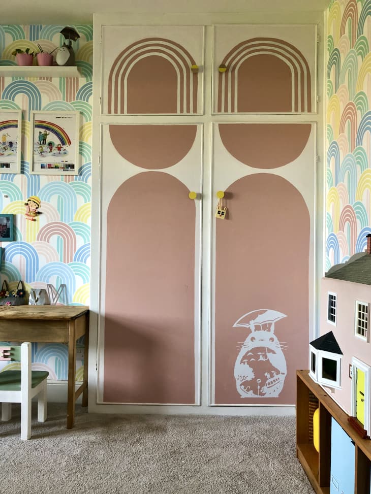 double closet doors, dusty rose curved mural painting on doors, rainbow wallpaper on walls, floor to ceiling closet doors, beige carpet, pink doll house, small wooden desk, white kids chair, floating shelf, kids art