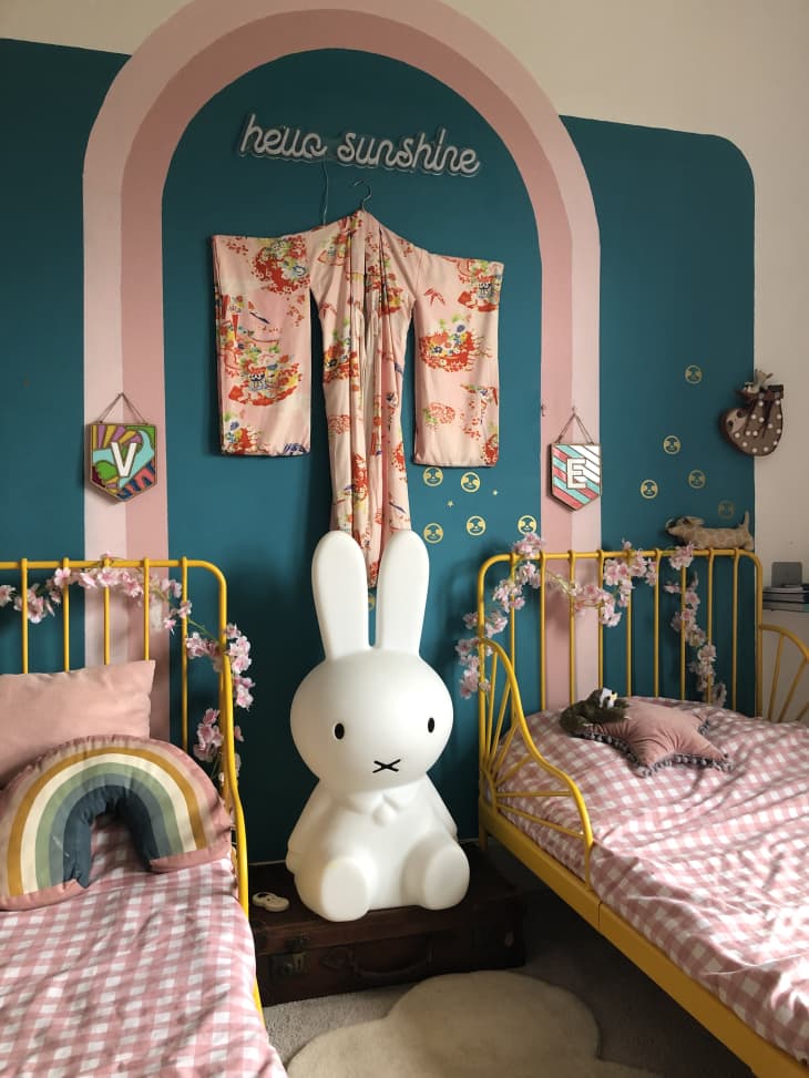 rainbow mural, blue walls painted 3/4 way up, life size light up bunny, two matching twin beds, yellow metal headboards, kimono on wall, hello sunshine neon sign, rainbow pillow, purple grid sheets, flower garland on headboards, v and e graphic flags