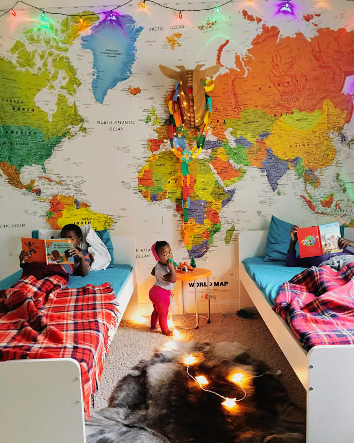 big colorful map, wall mural map, two twin white wood beds, plaid blankets in red and pink, colorful string lights, throw rug, small round orange side table, beige carpets