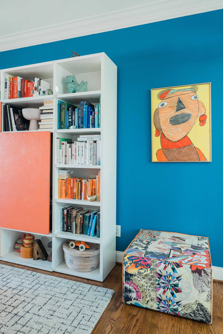 blue walls, color coded book shelf, colorful floor ottoman, orange painting, wood floors, white rug