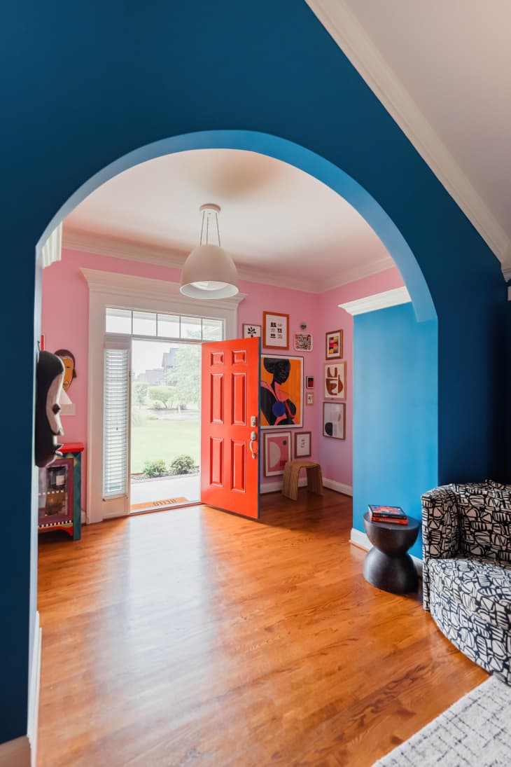pink walls, bold art, red door, foyer, large lighting fixture, wood floors, colorful hand painted cabinet, blue arch, mask on the wall, black and white patterned armchair