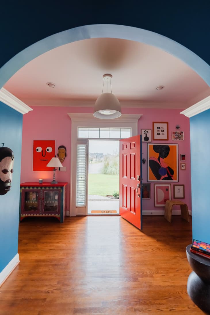 pink walls, bold art, red door, foyer, large lighting fixture, wood floors, colorful hand painted cabinet, blue arch, mask on the wall