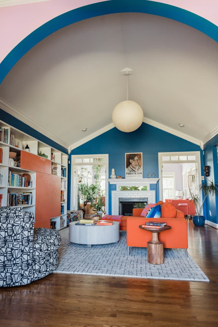 orange couch, black and white patterned chair, blue wall, vaulted ceiling, round chandelier, fireplace, color coded book shelf, orange painting, plants, round coffee table, living room, round wood end table, arched entry, bright sunlight