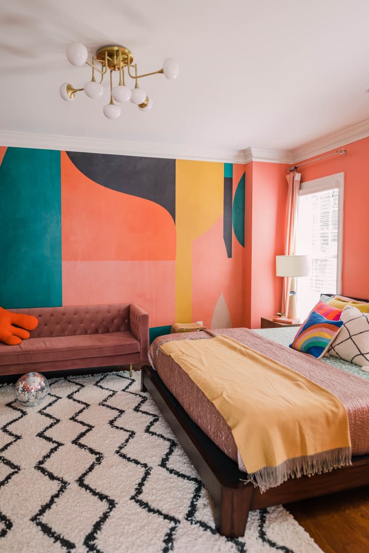 bedroom, colorful mural wall, pink walls, black and white textured rug, wood floors, yellow throw blanket, dusty pink couch, orb chandelier