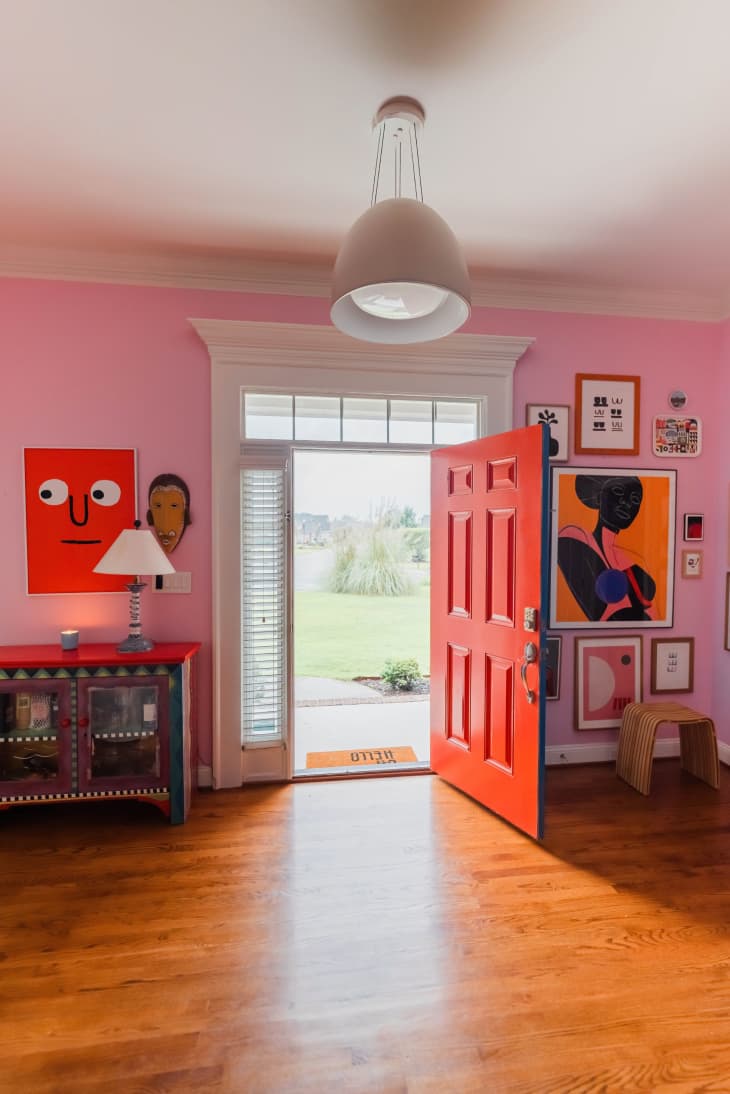 pink walls, bold art, red door, foyer, large lighting fixture, wood floors, colorful hand painted cabinet