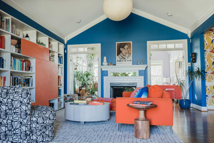 orange couch, black and white patterned chair, blue wall, vaulted ceiling, round chandelier, fireplace, color coded book shelf, orange painting, plants, round coffee table, living room, round wood end table