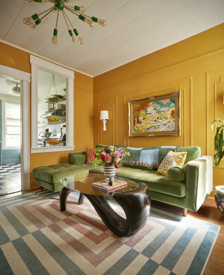 living room with yellow walls/wainscoting, green velvet sofa with colorful pillows, blue and green checkered throw, white wall sconces that are shaped like table lamps, artistic curved base coffee table with glass top, tree in colorful pot, painting with gold frame over sofa, yellow and green sputnik style ceiling light fixture