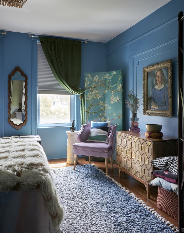Part of bedroom. Walls are blue with wainscoting, credenza with cool design, painting of woman with gold frame, velvet lilac armchair, blue screen with blossoming trees, blue shag rug on wood floor. foot of bed with faux fur throw visible