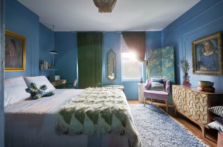 One wall of bedroom. Wall is blue with wainscoting, credenza with cool design, painting of woman with gold frame, velvet lilac armchair, blue screen with blossoming trees, blue shag rug on wood floor, bed has pink and pale blue bedding, faux fur throw. Large windows in back with green curtains