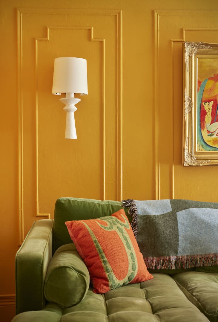 Corner of green velvet sofa in room with yellow walls with wainscoting. White wall sconce shaped like a table lamp