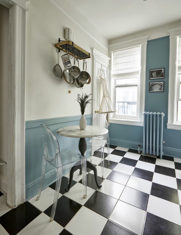 dining area in kitchen  with small round marble bistro table with clear lucite chairs. Black and white checkered floor, blue walls and radiator with white trim. Pots and pans hanging above dining table