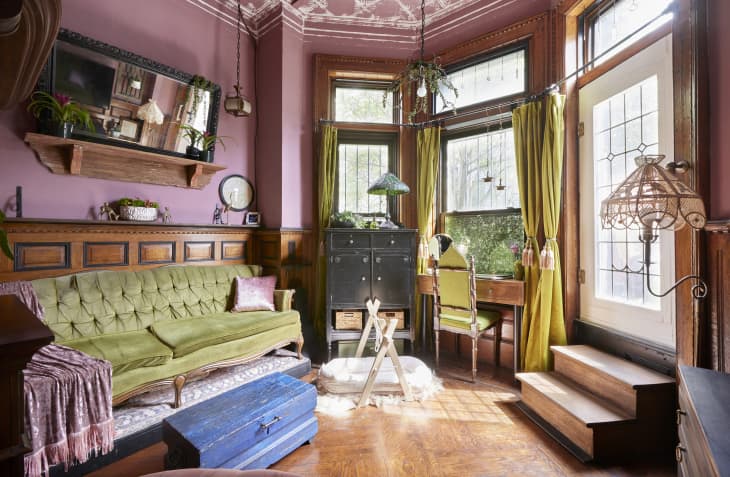 living room with pink walls, pink and white decorative ceiling, tall yellow/green curtains, green velvet antique sofa with shiny pink velvet pillow and throw, wood floors, 3 large windows, antique desk with pale green chair