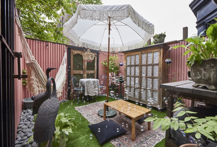 Patio with pink walls, fake grass, wood table with large white umbrella, wood and glass french doors, an off white hammock, bird statues, plants