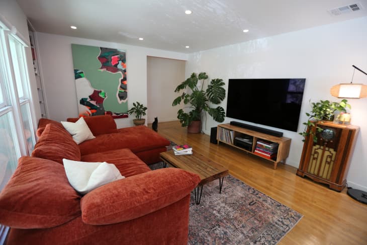 Rust colored sectional in plant filled  newly renovated living room with wooden coffee table across from media console. Colorful map painting hangs on wall near sofa.