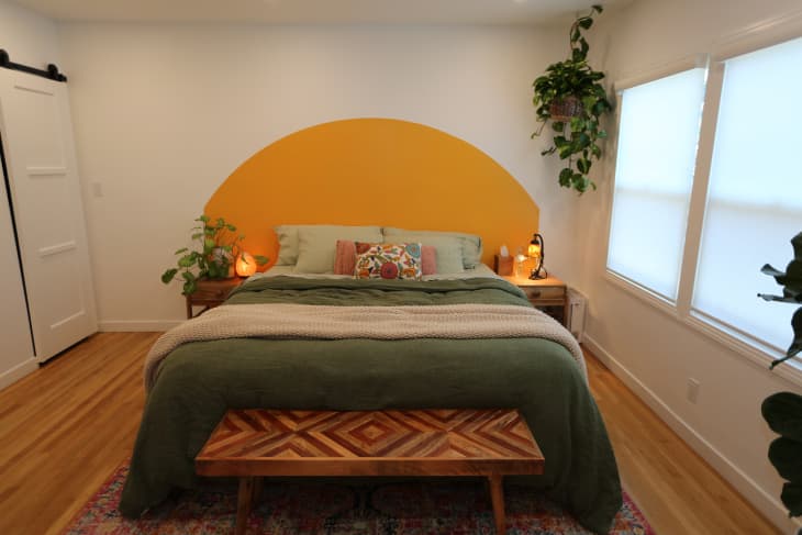 Semi-circular yellow wall decal behind neatly made bed with green bedding in newly renovated bedroom. Wooden bench sits at the end of the bed.