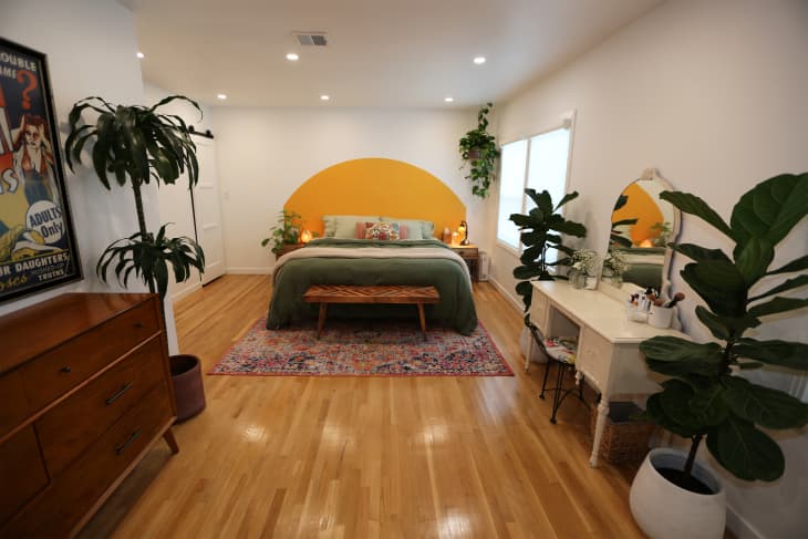 Semi-circular yellow wall decal sits behind neatly made bed with green bedding in newly renovated bedroom with white vanity desk across from wooden dresser.