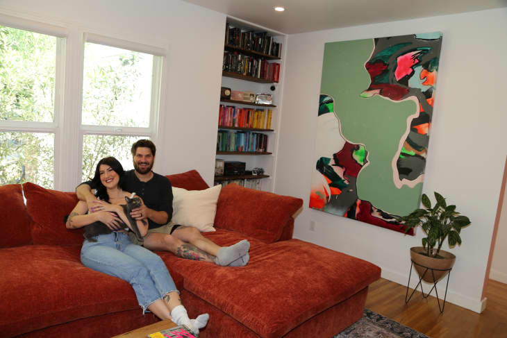 Couple holds cat while sitting on rust colored sofa in newly renovated living room with colorful map painting.