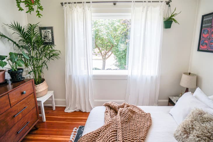 Various house plants in light filled bedroom with white linens on neatly made bed draped with taupe knitted blanket.