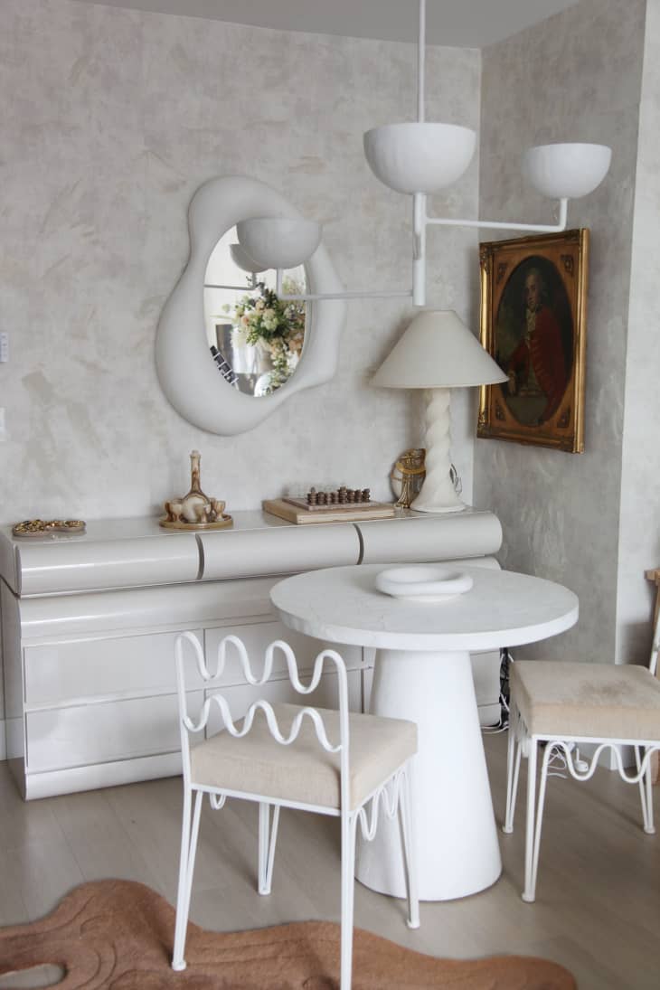 Dining area of apartment with small round bistro table and two squiggly dining chairs surrounding. Multi-armed pendant hangs above. Squiggly white mirror mounted on wall above white acrylic console.