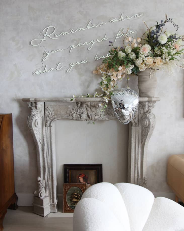 Fireplace mantel displayed on wall with large floral arrangement on top and deflated disco ball drooped over; neon signage above that reads "Remember when you prayed for what you have".